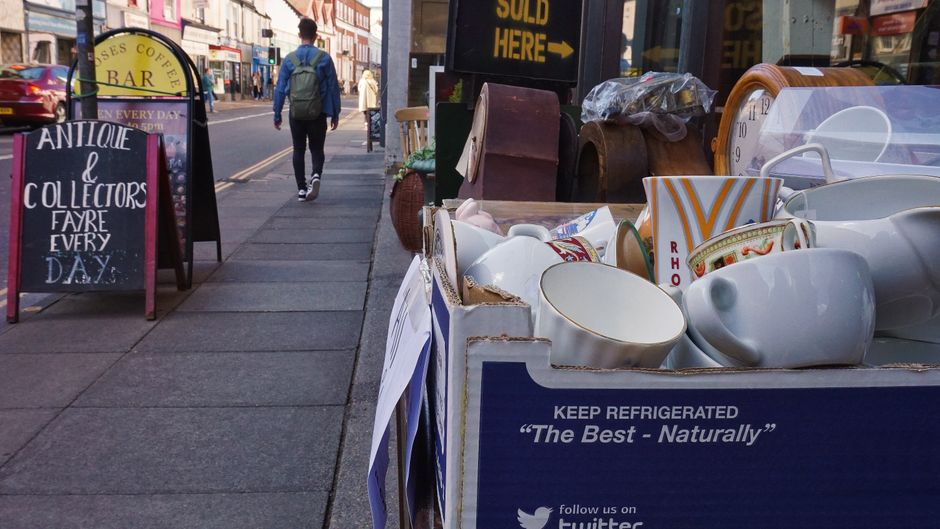 A street scene showing a sidewalk with a large cardboard box filled with white ceramic mugs and cups, some decorated with red and gold patterns. The box has a label reading 'KEEP REFRIGERATED' and 'Th