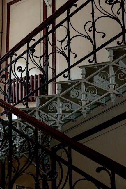 The image shows a narrow, steep staircase inside a property, featuring worn blue-painted wooden steps with peeling paint. To the left, there is a white wall with visible pipes and a small wall-mounted