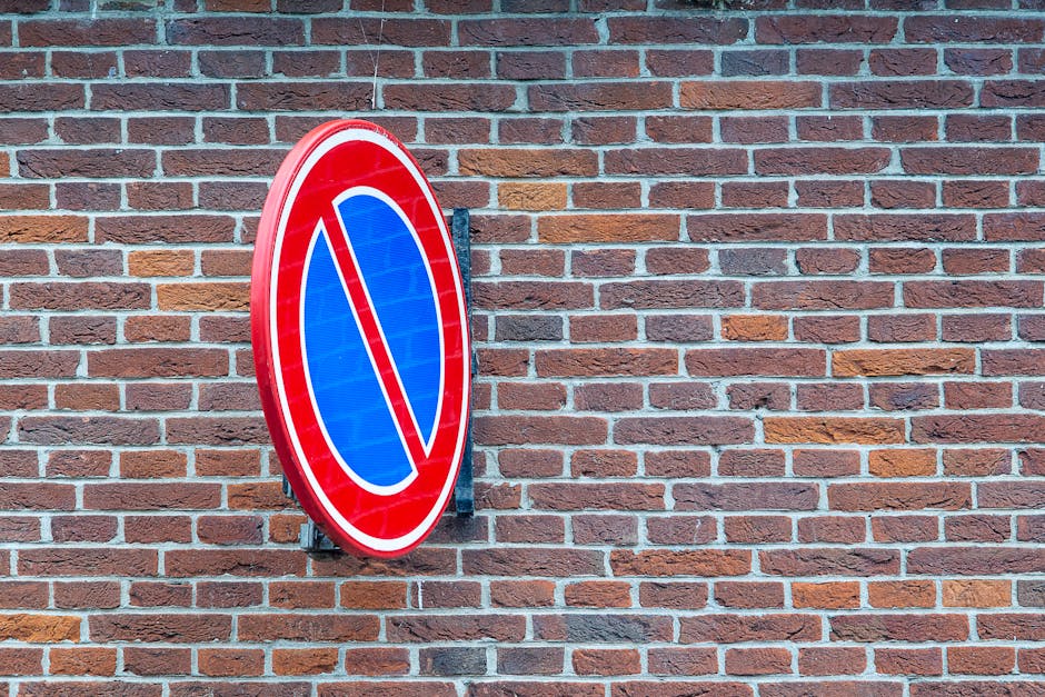 A round, blue and red no parking sign mounted on a black metal bracket attached to a red brick wall of a building. The brick wall features a traditional running bond pattern with reddish-brown and lig