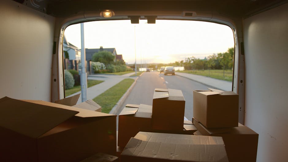 A vintage cream-colored van with a boxy design is pictured during daytime on a paved road with a metal guardrail on the side. The van is loaded with various furniture items, including wooden chairs an